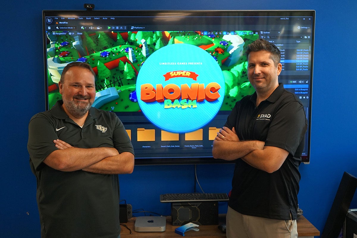 UCF Associate Professors Peter Smith and Matt Dombrowski stand in front of an screenshot of their new game Super Bionic Bash that is designed to help people learn to use their prosthetic limb.